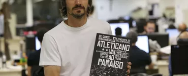 Com uma redação de jornal ao fundo e desfocada, Fred aparece em primeiro plano, de camiseta branca, cabelos castanhos lisos, na altura do queixo, bigode e barba rala, segurando o livro de sua autoria que tem a capa preta e branca, nas cores do Atlético.