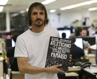 Com uma redação de jornal ao fundo e desfocada, Fred aparece em primeiro plano, de camiseta branca, cabelos castanhos lisos, na altura do queixo, bigode e barba rala, segurando o livro de sua autoria que tem a capa preta e branca, nas cores do Atlético.