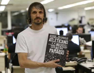 Com uma redação de jornal ao fundo e desfocada, Fred aparece em primeiro plano, de camiseta branca, cabelos castanhos lisos, na altura do queixo, bigode e barba rala, segurando o livro de sua autoria que tem a capa preta e branca, nas cores do Atlético.