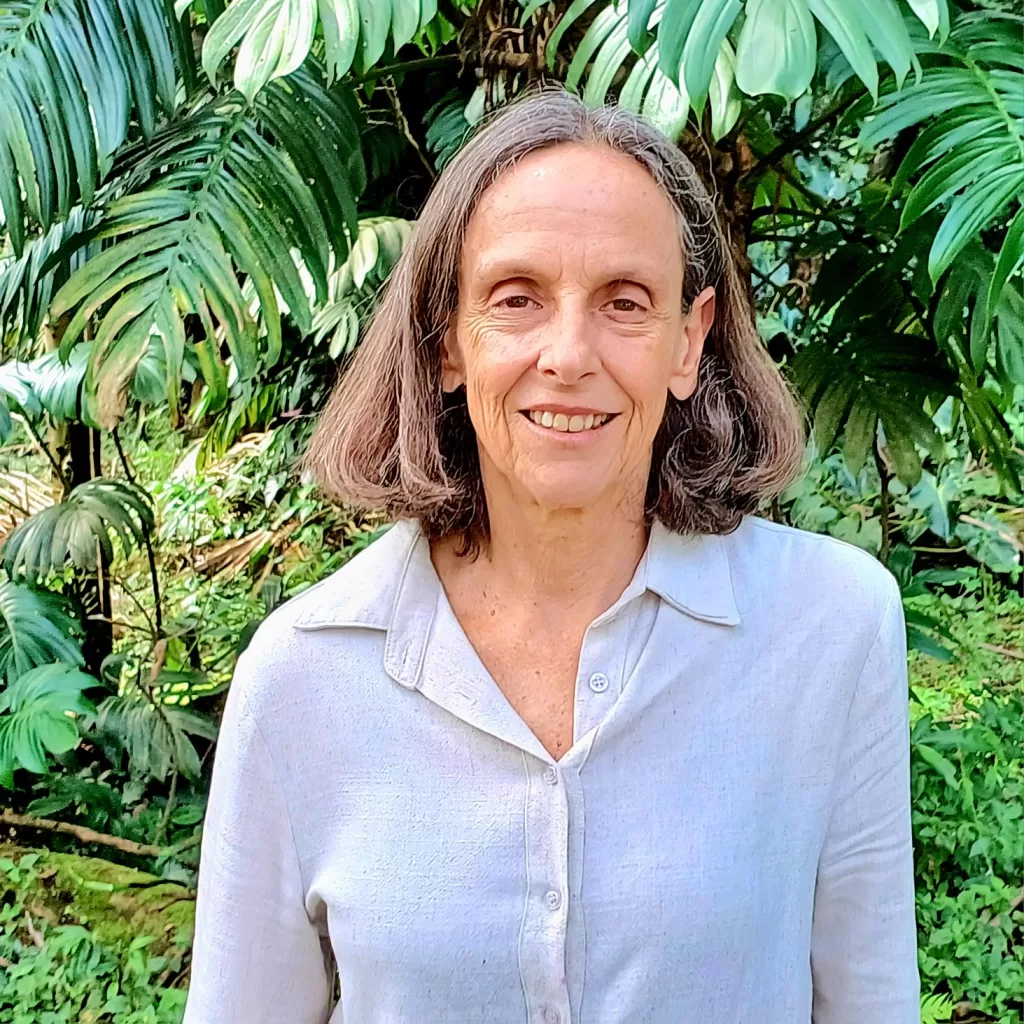 Maria é uma mulher branca, de cabelo curto grisalho; ela está em um ambiente aberto, em frente a plantas e veste uma camisa branca