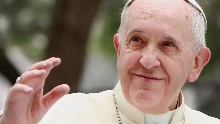 Papa Francisco em plano fechado sorri com os lábios, olha pra cima e expõe a mão como que para abençoar