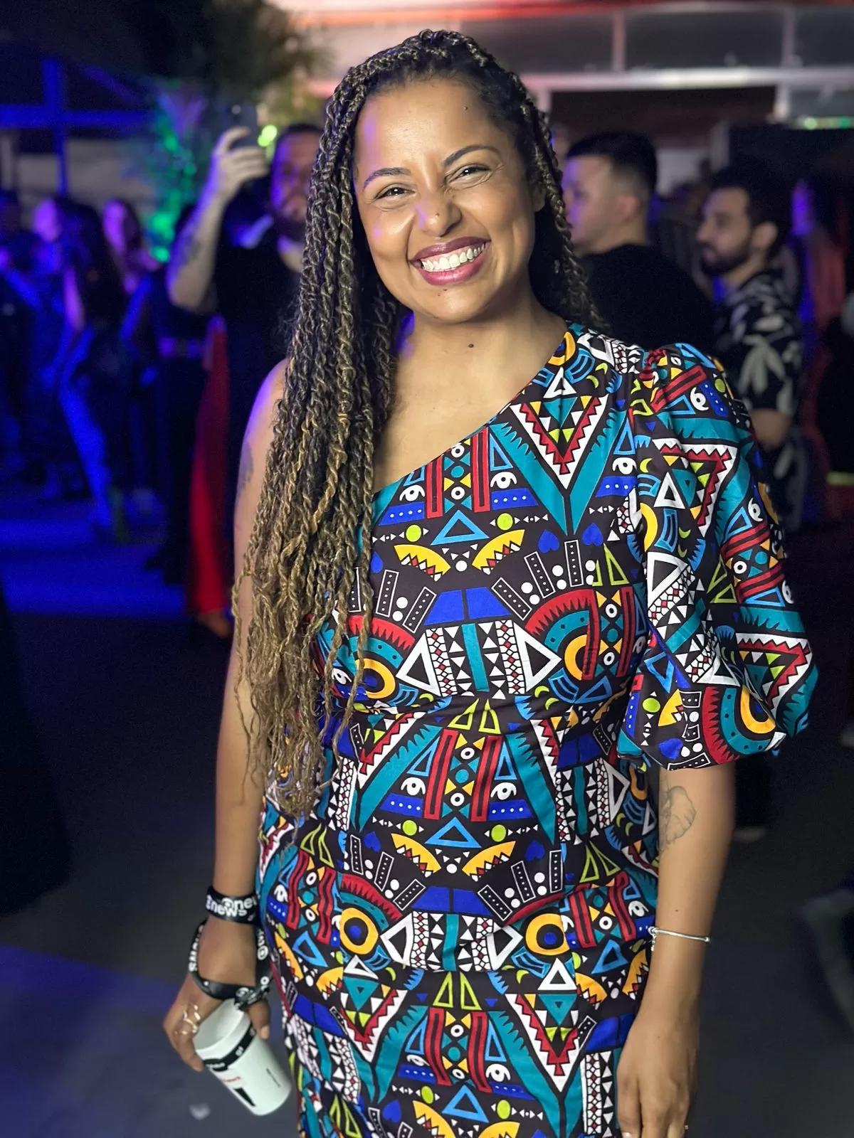 Tábata em fotografia em plano americano, sorrindo, em uma festa, segurando um copo. Ela é negra, está com o cabelo trançado e comprido, vestido colorido, de um ombro só, em estampa africana.