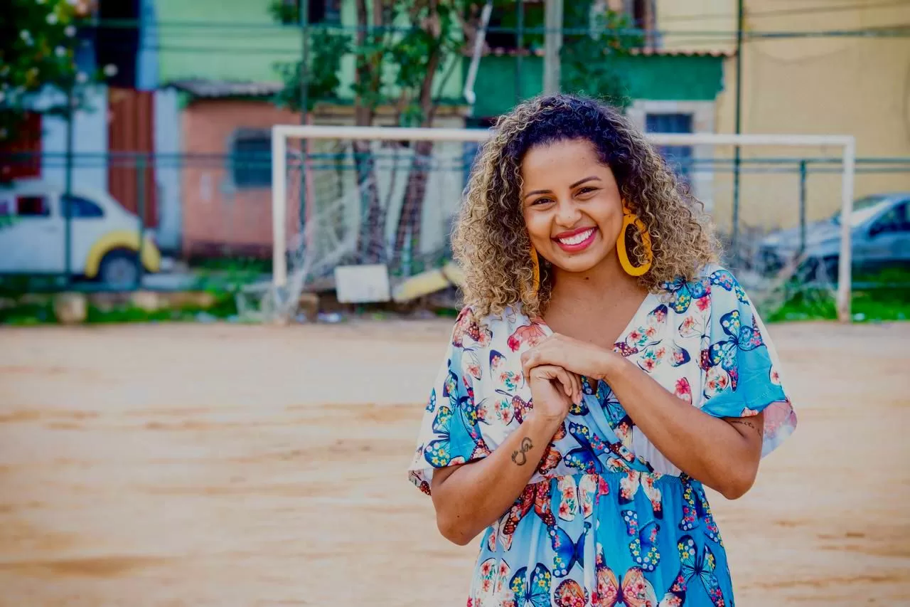 Tábata em primeiro plano, sorrindo em um campo de futebol de terra, com a trave do gol ao fundo. Ela tem o cabelo cacheado, curto e com luzes, com um vestido colorido com estampas de borboletas e brincos grandes laranjas.