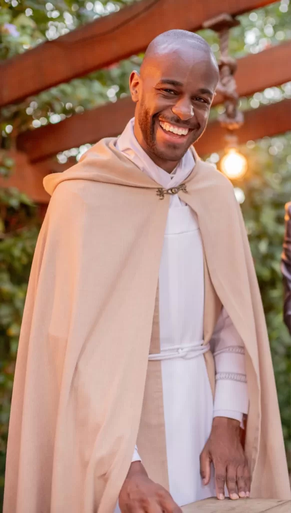 Samuel é um homem negro, careca de barba escura; ele usa uma túnica bege claro por cima de uma roupa religiosa branca. A imagem é vertical e mostra ele quase inteiro.