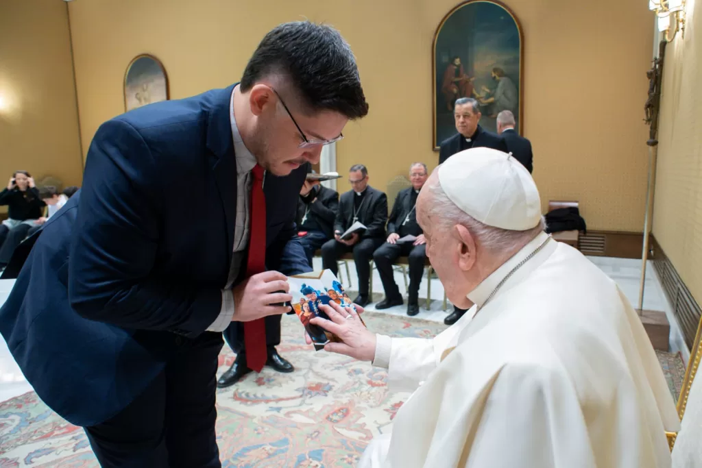 Marcus é um homem branco de cabelos escuros posicionado à esquerda da imagem, curvando-se diante do Papa Francisco, que está sentado à direita