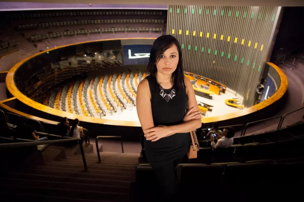 Foto de Carol Pires no início da carreira. Ela está em primeiro plano, em pé, com os braços cruzados e vestindo roupa preta, ao fundo o auditório do Congresso Nacional.