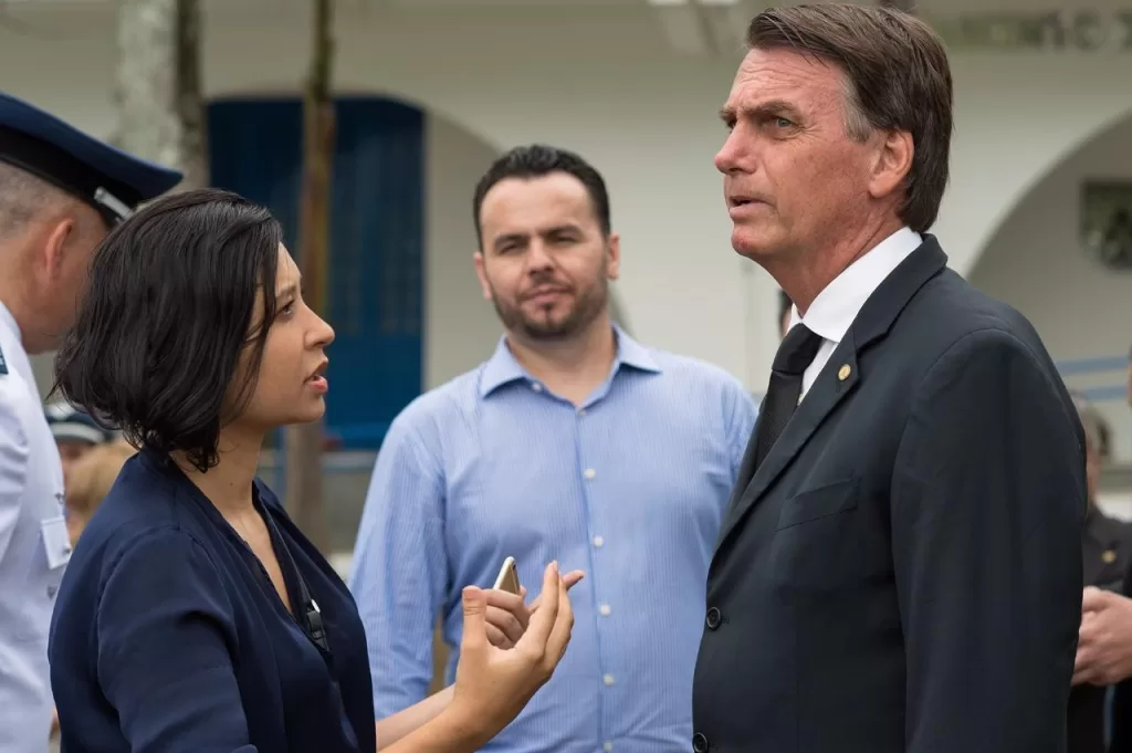 Carol Pires em conversa com o ex-presidente Jair Bolsonaro, ambos em pé, um de frente pro outro.