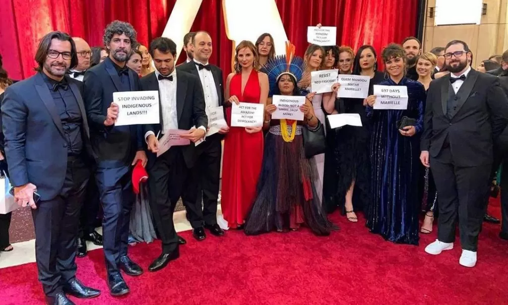 Equipe do documentário em pé no tapete vermelho do Oscar, segurando placas de protesto sobre temas políticos relevantes na época.