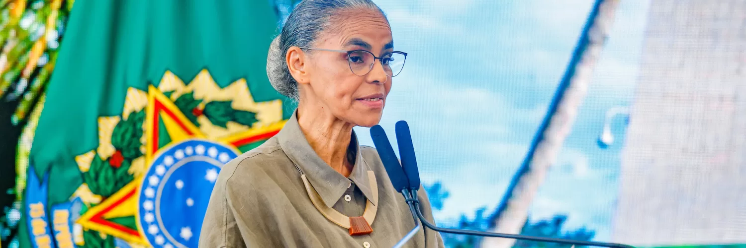 Marina Silva está no centro-esquerda da foto olhando para a direita com a bandeira com o brasão do Brasil no fundo