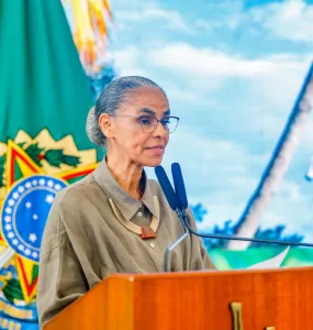 Marina Silva está no centro-esquerda da foto olhando para a direita com a bandeira com o brasão do Brasil no fundo