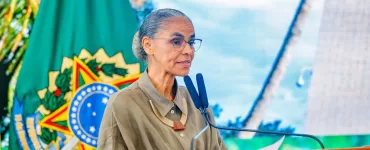 Marina Silva está no centro-esquerda da foto olhando para a direita com a bandeira com o brasão do Brasil no fundo