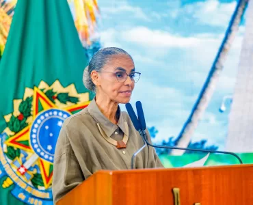 Marina Silva está no centro-esquerda da foto olhando para a direita com a bandeira com o brasão do Brasil no fundo