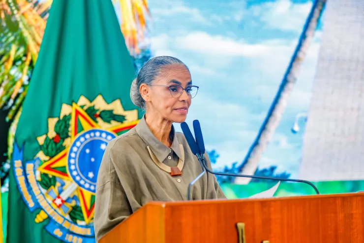 Marina Silva está no centro-esquerda da foto olhando para a direita com a bandeira com o brasão do Brasil no fundo