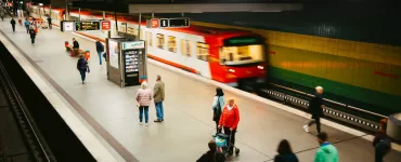 Metrô vermelho em movimento rápido passando por uma estação moderna com plataforma iluminada e poucos passageiros.