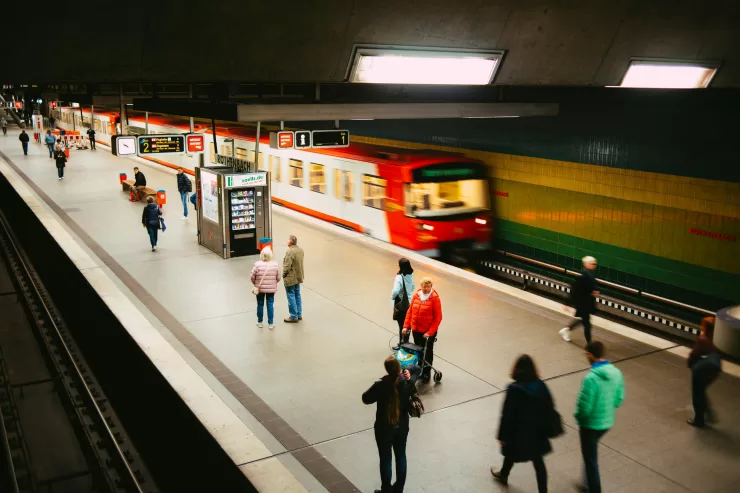 Metrô vermelho em movimento rápido passando por uma estação moderna com plataforma iluminada e poucos passageiros.