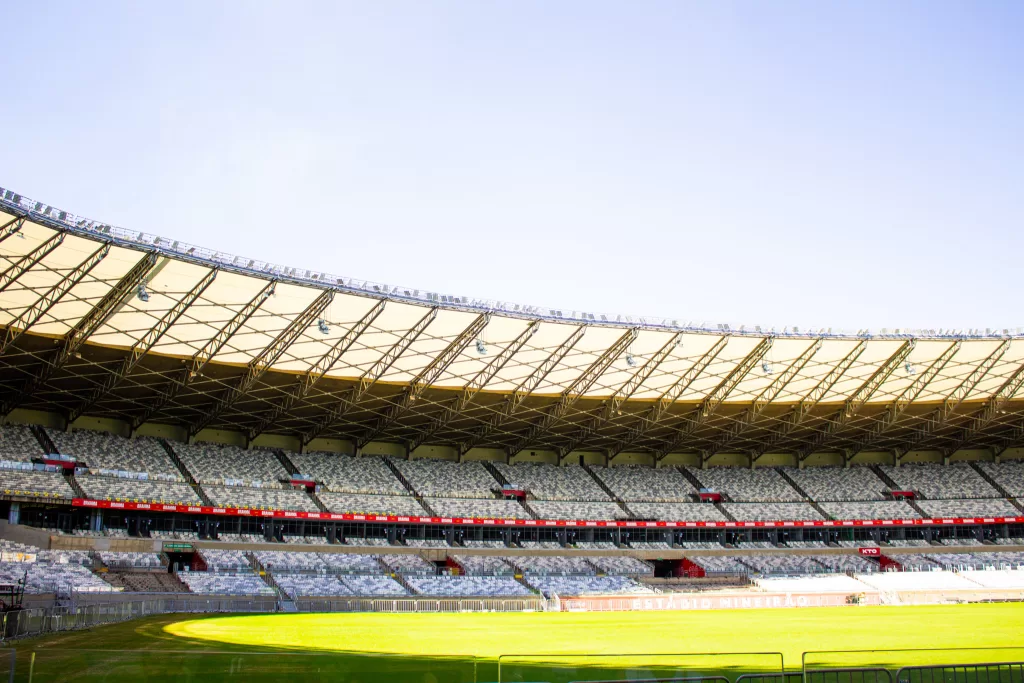 Foto da parte interna do estádio Mineirão, arquibancadas cinzas, estrutura de metal no alto, e no chão o gramado da cor verde sendo iluminado pelo sol.