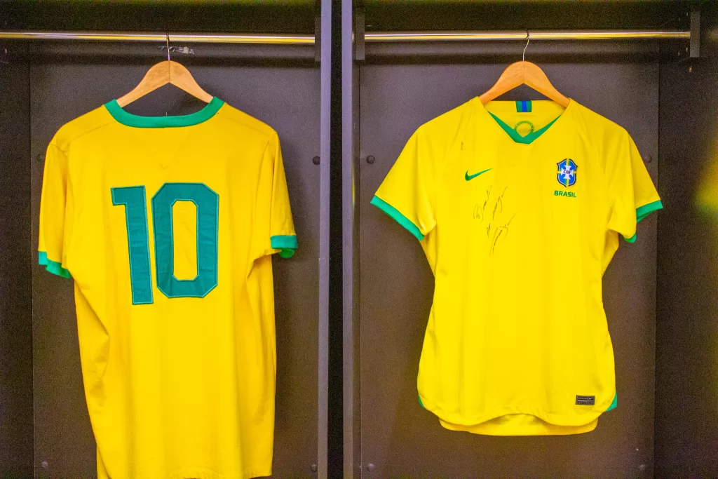 Duas camisas amarelas do Brasil penduradas em cabides de um armário. A esquerda, a parte de trás da camisa contém o número dez na cor verde, e na direita a parte de frente de uma camisa feminina, contém o brasão da CBF, o logo da Nike e um autógrafo não identificado.