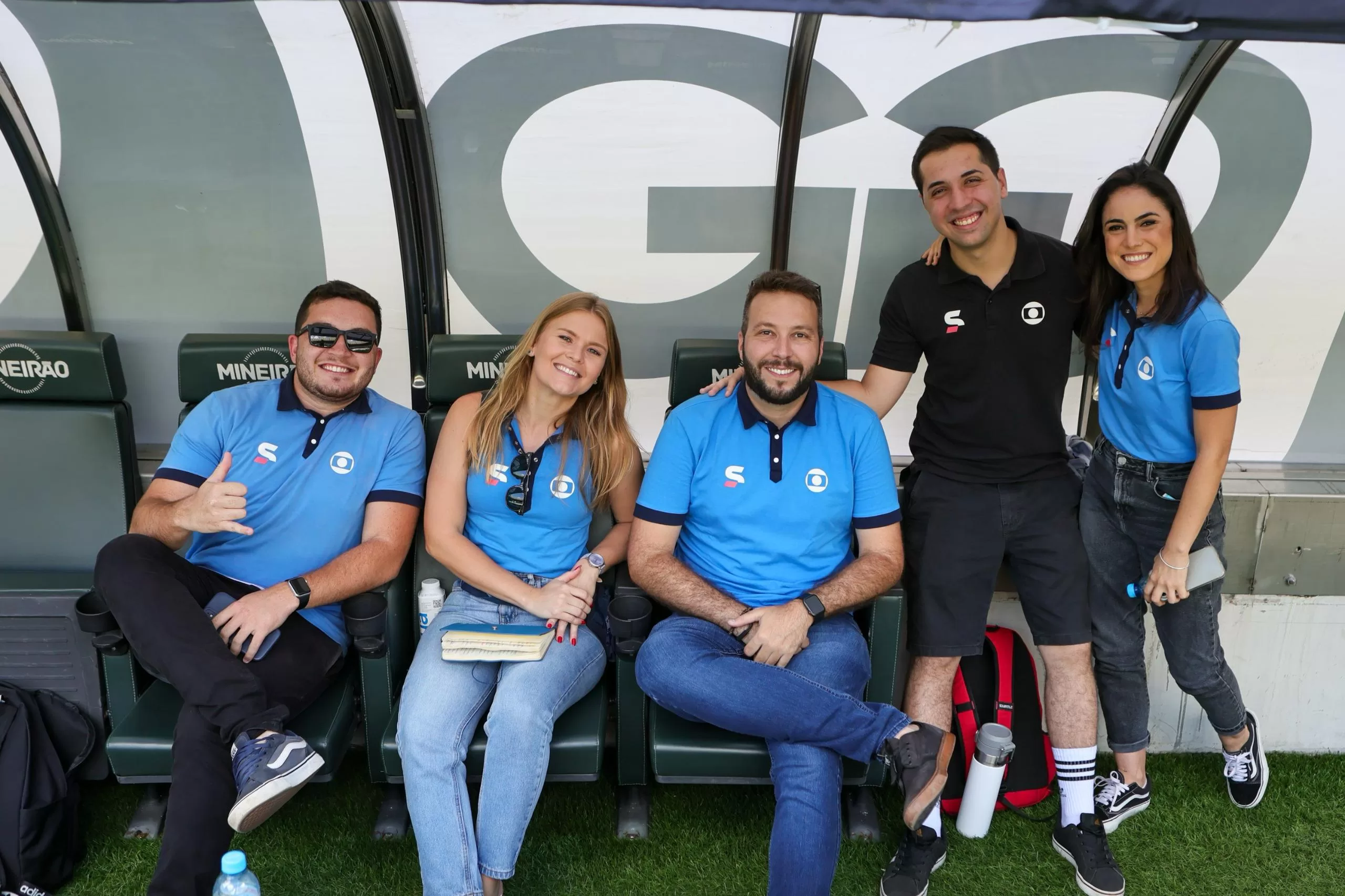 Cinco pessoas no banco de jogadores de um estádio de futebol. Três sentadas e duas pessoas em pé ao lado.