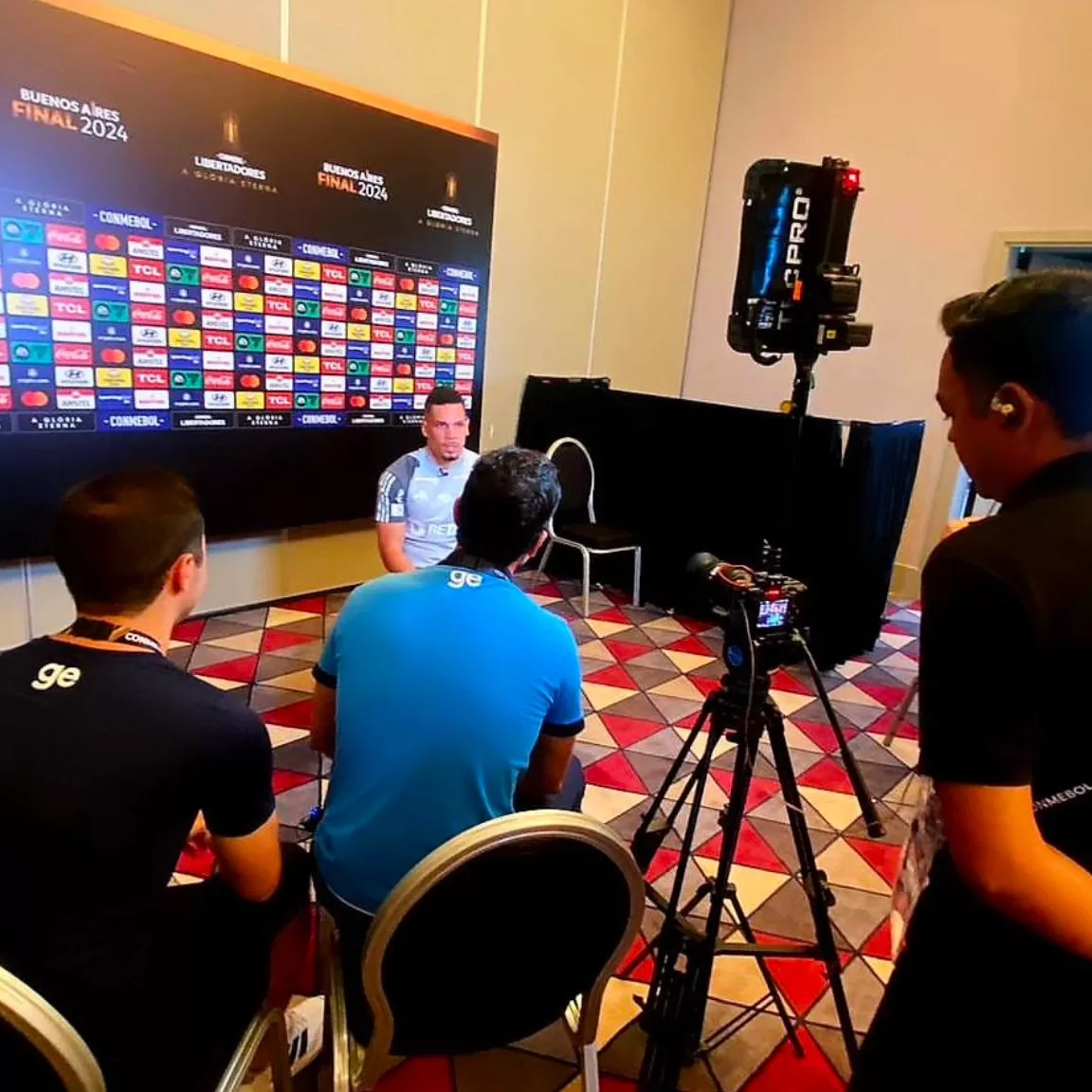Um homem de frente e três pessoas de costas durante uma entrevista. Um tripê com iluminação do lado direito da foto.