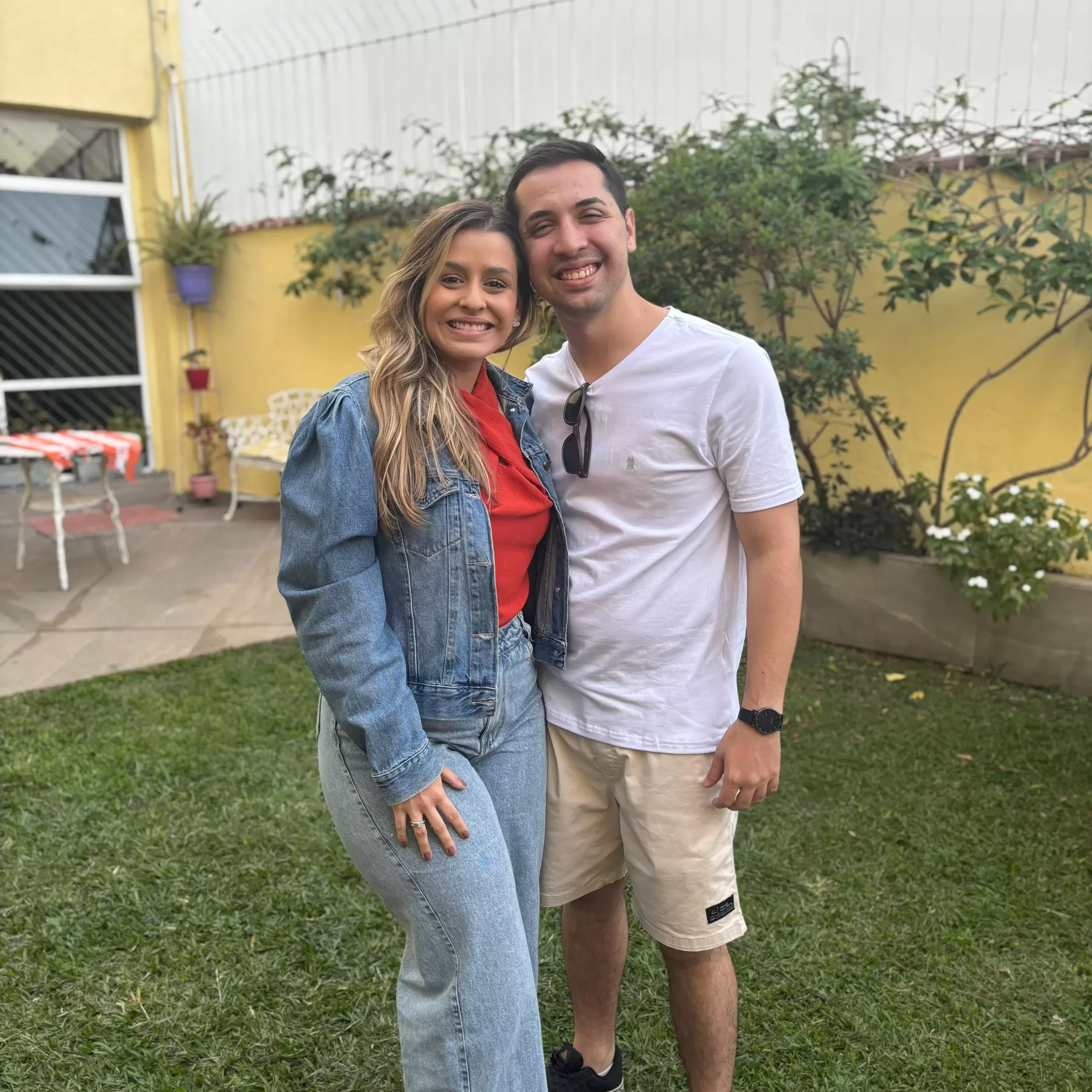 Um  homem abraçado de lado com uma mulher em um jardim. Ambos estão sorrindo.