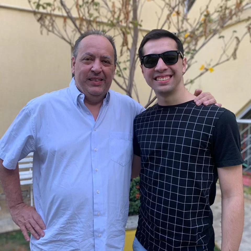 Dois homens em pé. O da esquerda está com a mão no ombro do homem da direita. Ambos estão sorrindo.