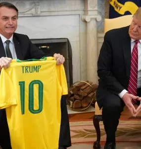Ex-presidente Jair Bolsonaro sentado ao lado do presidente dos Estados Unidos Donald Trump. Bolsonaro sorri enquanto segura uma camisa amarela da seleção brasileira de futebol com o nome “Trump” escrito.
