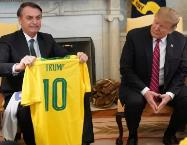 Ex-presidente Jair Bolsonaro sentado ao lado do presidente dos Estados Unidos Donald Trump. Bolsonaro sorri enquanto segura uma camisa amarela da seleção brasileira de futebol com o nome “Trump” escrito.