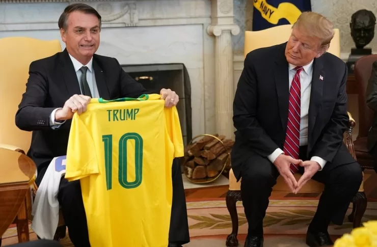 Ex-presidente Jair Bolsonaro sentado ao lado do presidente dos Estados Unidos Donald Trump. Bolsonaro sorri enquanto segura uma camisa amarela da seleção brasileira de futebol com o nome “Trump” escrito.