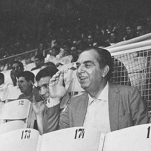 Fotografia de Nelson Rodrigues em primeiro plano em preto e branco, acenando e sorrindo, em arquibancada de um estádio de futebol. Está vestido com roupa formal, camisa social e paletó, com pessoas à sua volta. 
