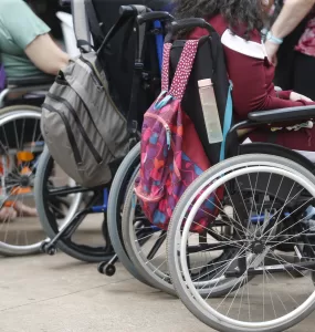 Pessoas cadeirantes estão enfileiradas, com mochilas penduradas na parte de trás das cadeiras de rodas.