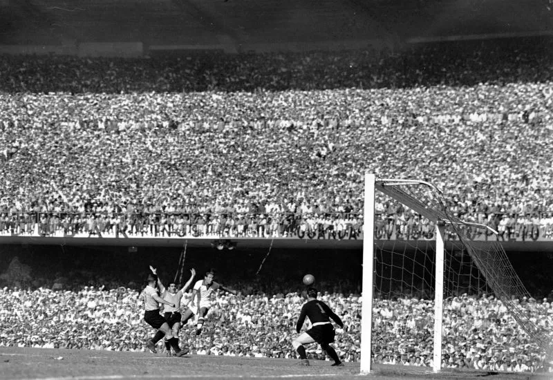 Fotografia em preto e branco de partida de futebol, Brasil contra Uruguai, no estádio Maracanã. Em primeiro plano a trave do gol, três jogadores correndo, a bola no ar e o goleiro olhando em direção à bola. A Geral e o Anel Superior estão lotados com torcedores