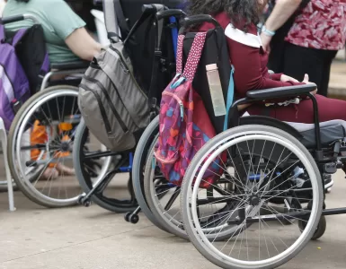 Pessoas cadeirantes estão enfileiradas, com mochilas penduradas na parte de trás das cadeiras de rodas.