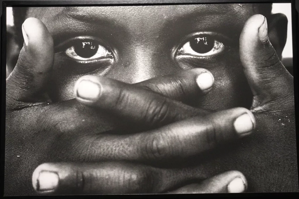 Fotografia em preto e branco com criança negra em primeiro plano, cobrindo a boca e o nariz com as mãos entrelaçadas. 
