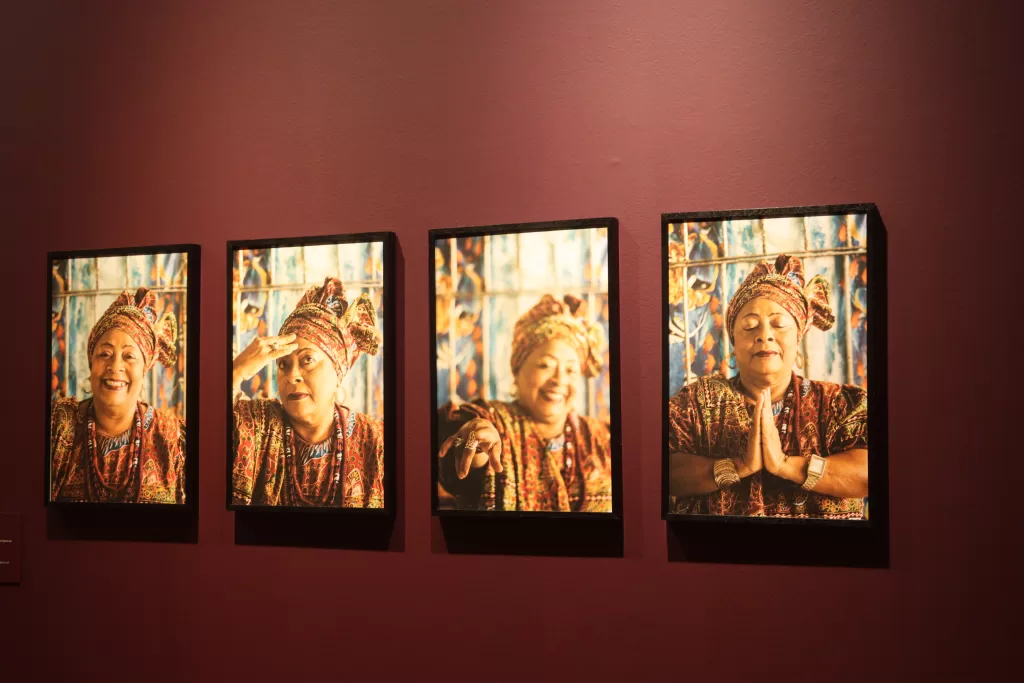 Quatro quadros de fotografias enfileiradas em primeiro plano, pendurados em uma parede marrom. As fotografias são compostas por uma mulher de idade, vestida com roupa de baiana e turbante na cabeça, fazendo poses diferentes.  
