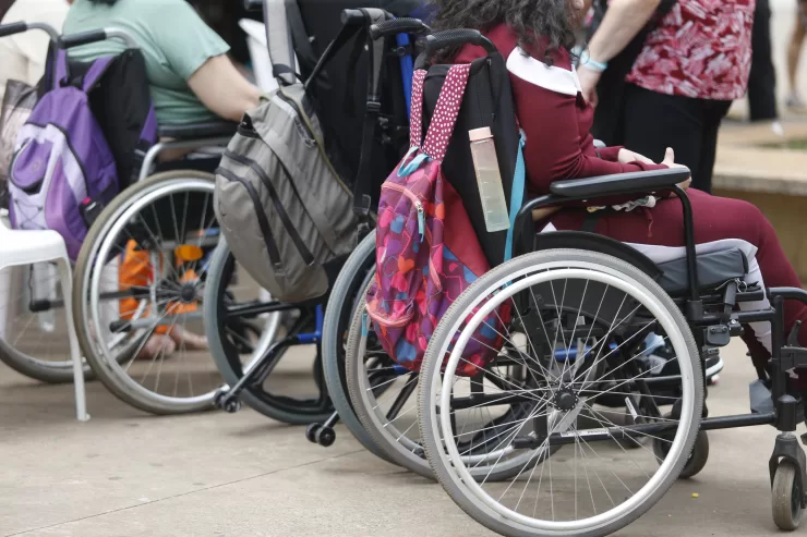 Pessoas cadeirantes estão enfileiradas, com mochilas penduradas na parte de trás das cadeiras de rodas.