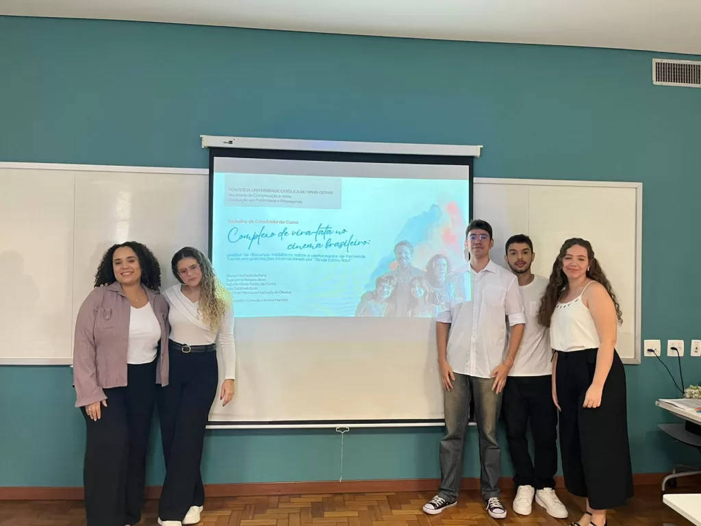 Alunos na sala de aula de frente à um quadro branco, sendo projetado o trabalho com o título. Estão vestidos com roupas formais.
