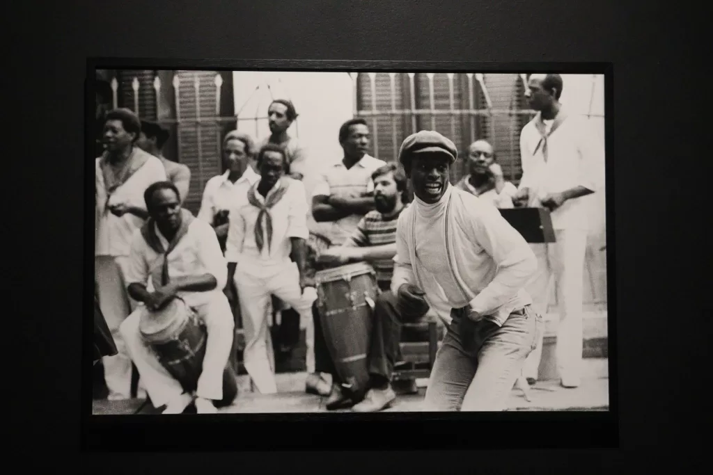 Fotografia em preto e branco, com um grupo de pessoas dançando e tocando o instrumento surdo. O grupo é composto por pessoas majoritariamente negras, que usam roupas claras e estão na rua de frente para uma casa. 
