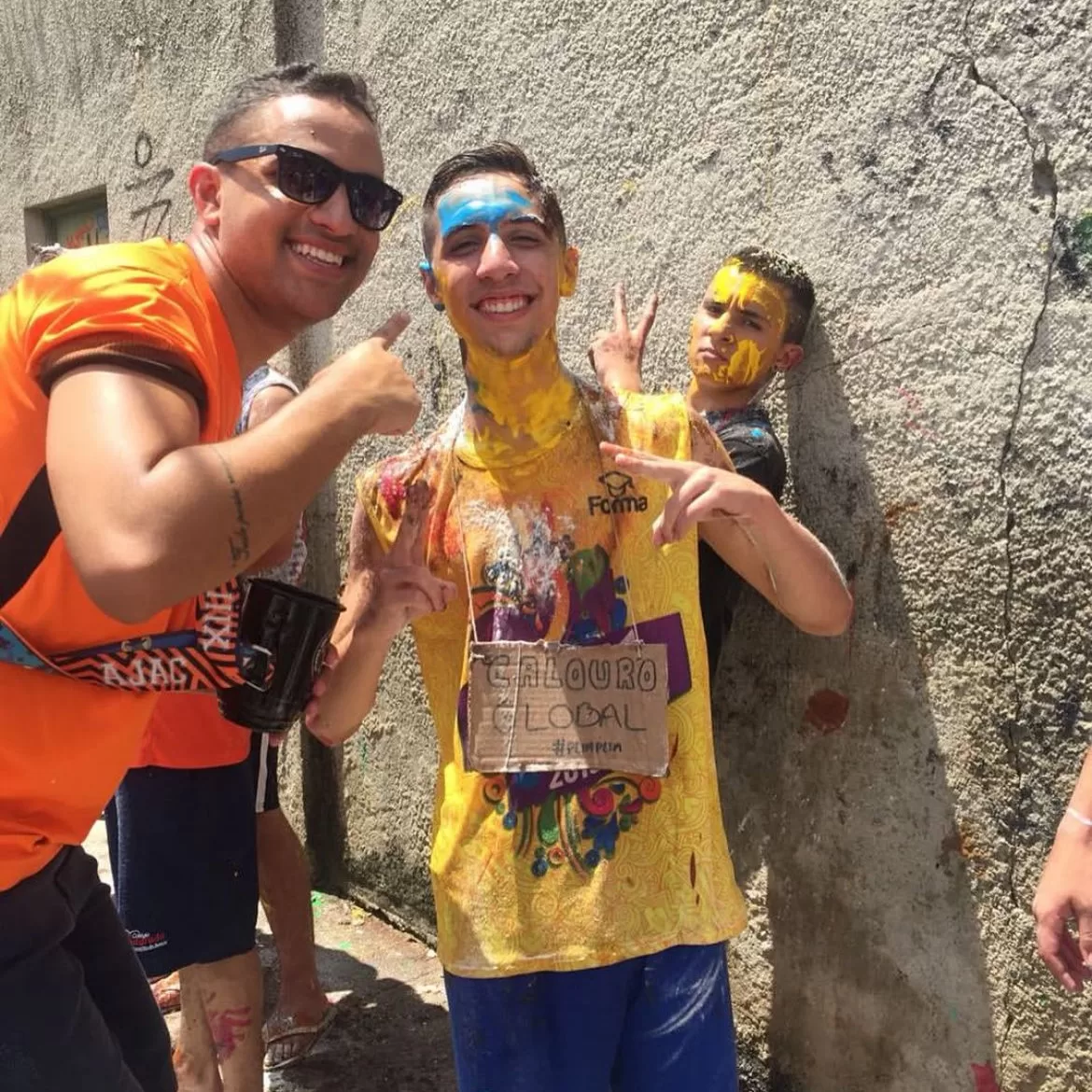 Pedro Spinelli sorrindo no trote de calouros da faculdade sujo de tinta. No pescoço, uma placa de papelão pendurada escrito "Calouro global #plimplim". Ao seu lado, um homem apontando para ele.