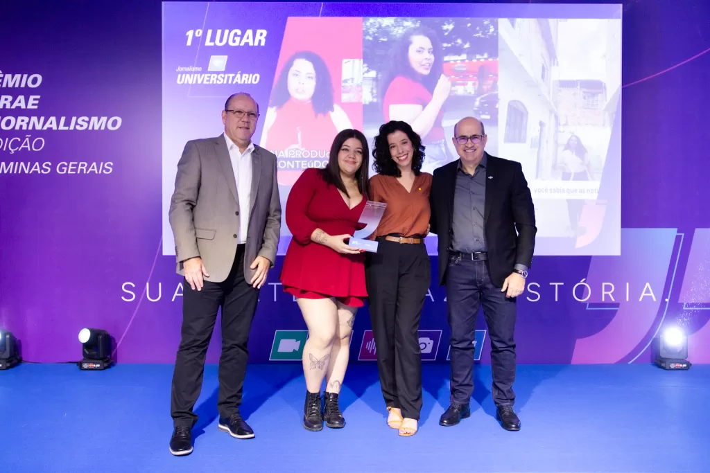 A imagem mostra quatro pessoas adultas em pé, sorrindo e posando para uma foto em um palco, com um telão digital ao fundo. A cena é de uma cerimônia de premiação noturna, com iluminação de palco na cor azul. No centro, duas mulheres, uma vestindo um vestido vermelho e a outra uma blusa marrom, seguram um troféu de acrílico transparente. À esquerda da imagem, um homem de terno cinza e calça preta, e à direita, um homem de blazer preto e jeans escuro. O telão digital ao fundo exibe a frase "1º LUGAR" e imagens de pessoas em um contexto de reportagem.