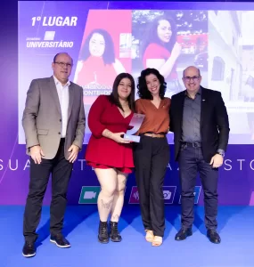 A imagem mostra quatro pessoas adultas em pé, sorrindo e posando para uma foto em um palco, com um telão digital ao fundo. A cena é de uma cerimônia de premiação noturna, com iluminação de palco na cor azul. No centro, duas mulheres, uma vestindo um vestido vermelho e a outra uma blusa marrom, seguram um troféu de acrílico transparente. À esquerda da imagem, um homem de terno cinza e calça preta, e à direita, um homem de blazer preto e jeans escuro. O telão digital ao fundo exibe a frase "1º LUGAR" e imagens de pessoas em um contexto de reportagem.