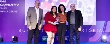 A imagem mostra quatro pessoas adultas em pé, sorrindo e posando para uma foto em um palco, com um telão digital ao fundo. A cena é de uma cerimônia de premiação noturna, com iluminação de palco na cor azul. No centro, duas mulheres, uma vestindo um vestido vermelho e a outra uma blusa marrom, seguram um troféu de acrílico transparente. À esquerda da imagem, um homem de terno cinza e calça preta, e à direita, um homem de blazer preto e jeans escuro. O telão digital ao fundo exibe a frase "1º LUGAR" e imagens de pessoas em um contexto de reportagem.