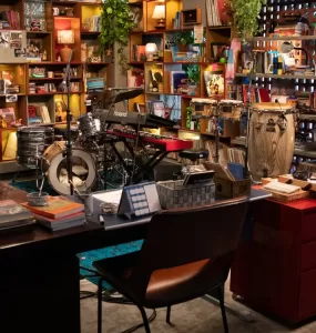 Uma vista panorâmica de um estúdio ou escritório aconchegante e eclético, com prateleiras de madeira repletas de livros, discos de vinil, plantas e diversos objetos decorativos. No centro, há uma bateria, um teclado e percussão dispostos em um palco. Uma mesa de escritório com livros e canecas está em primeiro plano, com uma cadeira marrom à frente dela. A iluminação é quente e convidativa.