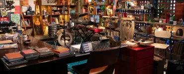 Uma vista panorâmica de um estúdio ou escritório aconchegante e eclético, com prateleiras de madeira repletas de livros, discos de vinil, plantas e diversos objetos decorativos. No centro, há uma bateria, um teclado e percussão dispostos em um palco. Uma mesa de escritório com livros e canecas está em primeiro plano, com uma cadeira marrom à frente dela. A iluminação é quente e convidativa.