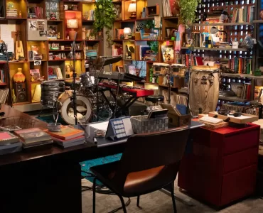 Uma vista panorâmica de um estúdio ou escritório aconchegante e eclético, com prateleiras de madeira repletas de livros, discos de vinil, plantas e diversos objetos decorativos. No centro, há uma bateria, um teclado e percussão dispostos em um palco. Uma mesa de escritório com livros e canecas está em primeiro plano, com uma cadeira marrom à frente dela. A iluminação é quente e convidativa.