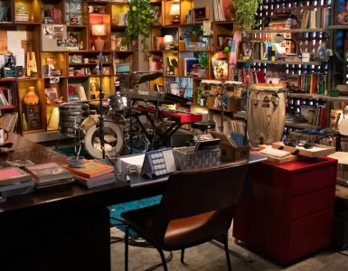 Uma vista panorâmica de um estúdio ou escritório aconchegante e eclético, com prateleiras de madeira repletas de livros, discos de vinil, plantas e diversos objetos decorativos. No centro, há uma bateria, um teclado e percussão dispostos em um palco. Uma mesa de escritório com livros e canecas está em primeiro plano, com uma cadeira marrom à frente dela. A iluminação é quente e convidativa.