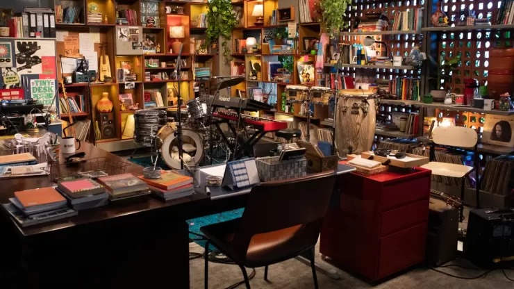 Uma vista panorâmica de um estúdio ou escritório aconchegante e eclético, com prateleiras de madeira repletas de livros, discos de vinil, plantas e diversos objetos decorativos. No centro, há uma bateria, um teclado e percussão dispostos em um palco. Uma mesa de escritório com livros e canecas está em primeiro plano, com uma cadeira marrom à frente dela. A iluminação é quente e convidativa.