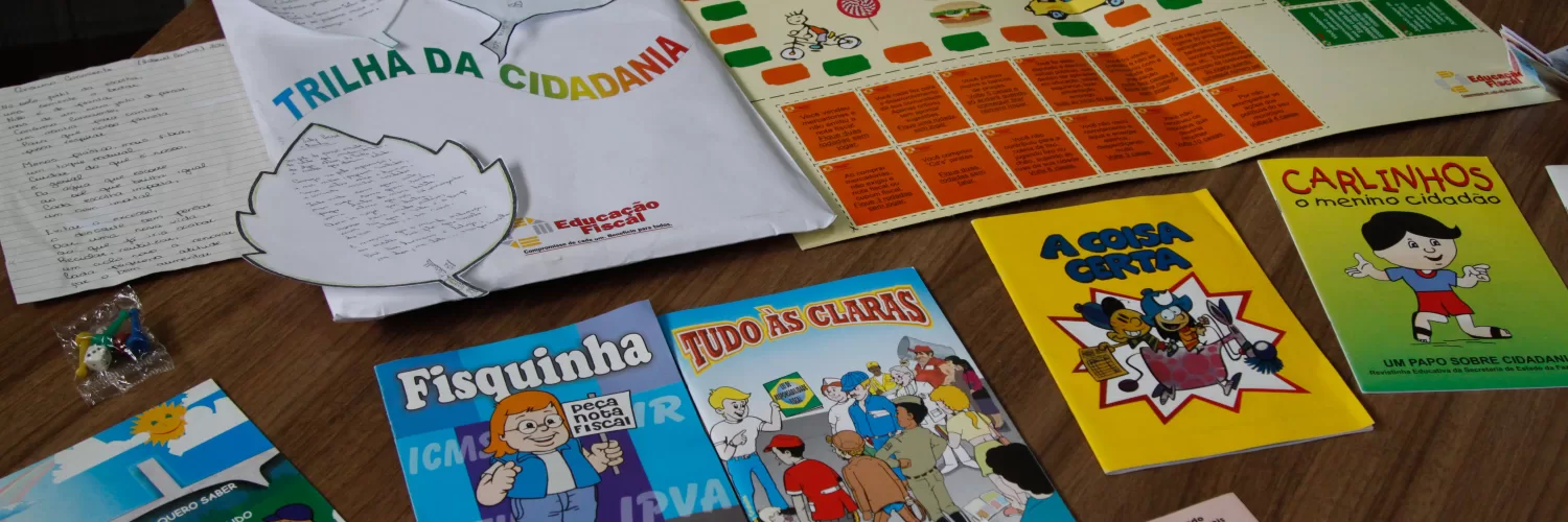 Materiais didáticos do Programa de Educação Fiscal de Minas Gerais estão dispostos sobre uma mesa de madeira. Entre eles, há cartilhas ilustradas com linguagem infantil, como “Fisquinha em ação”, “Tudo às claras”, “A coisa certa”, “Carlinhos, o menino cidadão” e “O menino quero saber”, além de folhetos explicativos sobre finanças públicas e cidadania. Também estão presentes jogos educativos, como o tabuleiro “Trilha da Cidadania”, utilizado para ensinar de forma lúdica o papel dos tributos e a importância da participação social. Os materiais são utilizados em escolas públicas para promover a Educação Fiscal entre estudantes da rede estadual.