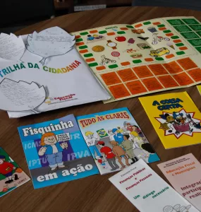 Materiais didáticos do Programa de Educação Fiscal de Minas Gerais estão dispostos sobre uma mesa de madeira. Entre eles, há cartilhas ilustradas com linguagem infantil, como “Fisquinha em ação”, “Tudo às claras”, “A coisa certa”, “Carlinhos, o menino cidadão” e “O menino quero saber”, além de folhetos explicativos sobre finanças públicas e cidadania. Também estão presentes jogos educativos, como o tabuleiro “Trilha da Cidadania”, utilizado para ensinar de forma lúdica o papel dos tributos e a importância da participação social. Os materiais são utilizados em escolas públicas para promover a Educação Fiscal entre estudantes da rede estadual.