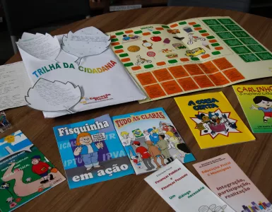 Materiais didáticos do Programa de Educação Fiscal de Minas Gerais estão dispostos sobre uma mesa de madeira. Entre eles, há cartilhas ilustradas com linguagem infantil, como “Fisquinha em ação”, “Tudo às claras”, “A coisa certa”, “Carlinhos, o menino cidadão” e “O menino quero saber”, além de folhetos explicativos sobre finanças públicas e cidadania. Também estão presentes jogos educativos, como o tabuleiro “Trilha da Cidadania”, utilizado para ensinar de forma lúdica o papel dos tributos e a importância da participação social. Os materiais são utilizados em escolas públicas para promover a Educação Fiscal entre estudantes da rede estadual.