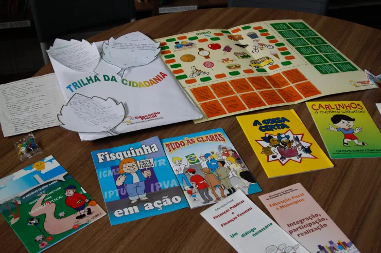 Materiais didáticos do Programa de Educação Fiscal de Minas Gerais estão dispostos sobre uma mesa de madeira. Entre eles, há cartilhas ilustradas com linguagem infantil, como “Fisquinha em ação”, “Tudo às claras”, “A coisa certa”, “Carlinhos, o menino cidadão” e “O menino quero saber”, além de folhetos explicativos sobre finanças públicas e cidadania. Também estão presentes jogos educativos, como o tabuleiro “Trilha da Cidadania”, utilizado para ensinar de forma lúdica o papel dos tributos e a importância da participação social. Os materiais são utilizados em escolas públicas para promover a Educação Fiscal entre estudantes da rede estadual.